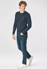 Maglione a coste navy con motivo a trecce, colletto e polsini a costine, abbinato a jeans blu scuro e sneakers bianche. Dettaglio del logo semplice sul maglione.