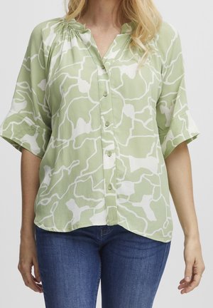 Žena nosí světle zelenou a bílou abstraktně vzorovanou košili na knoflíky s ohrnutými polovičními rukávy a modré džíny.