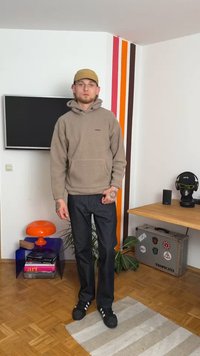 Hoodie de fleece bege com um bolso frontal, jeans de ganga escura e ténis pretos. Ambiente interior com riscas coloridas nas paredes e decoração.