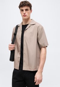 Jeune homme portant une chemise beige à manches courtes boutonnée par-dessus un t-shirt noir, un pantalon noir, et une sangle de sac épaule noire.