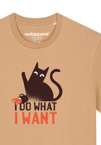 Beige Baumwoll-T-Shirt mit einer Illustration einer schwarzen Katze mit weißen Augen und dem Text "ICH MACH, WAS ICH WILL" in fetter orange- und schwarzer Schrift.