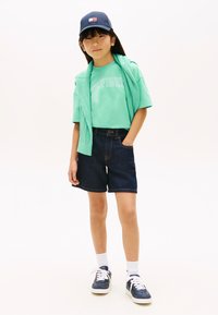 Πράσινο πουκάμισο με κοντά μανίκια και το κείμενο «Tommy», μπλε σκουφάκι, ελαφριά μπλούζα περασμένη πάνω από τους ώμους, σκουρόχρωμα σορτς τζιν, λευκές κάλτσες, μπλε αθλητικά παπούτσια.