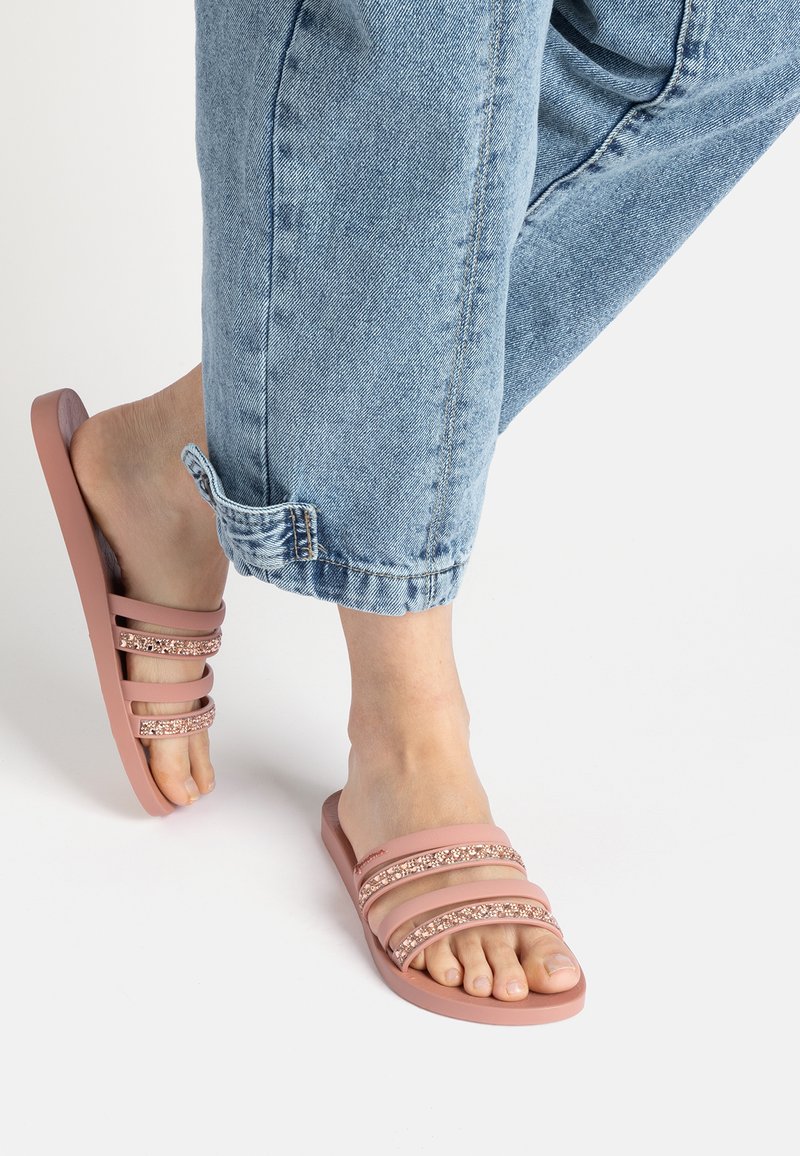 Rosa Rutsch-Sandalen mit drei breiten Riemen, von denen einer mit einem geschmückten Akzent verziert ist. Weiches Material mit flacher Sohle. Getragen mit hochgekrempelten Jeans.