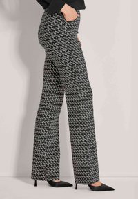 Vrouw in zwarte en witte geometrische patroon met wijde pijpen en zwarte puntige hoge hakken, zijwaarts poserend met hand in de zak.