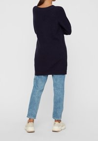 Tummansininen neulepusero rentoa leikkausta, joissa on ribbauskohdistuksia hihoissa, yhdistettynä vaaleansinisiin farkkuihin ja neutraalinsävyisiin tennareihin.