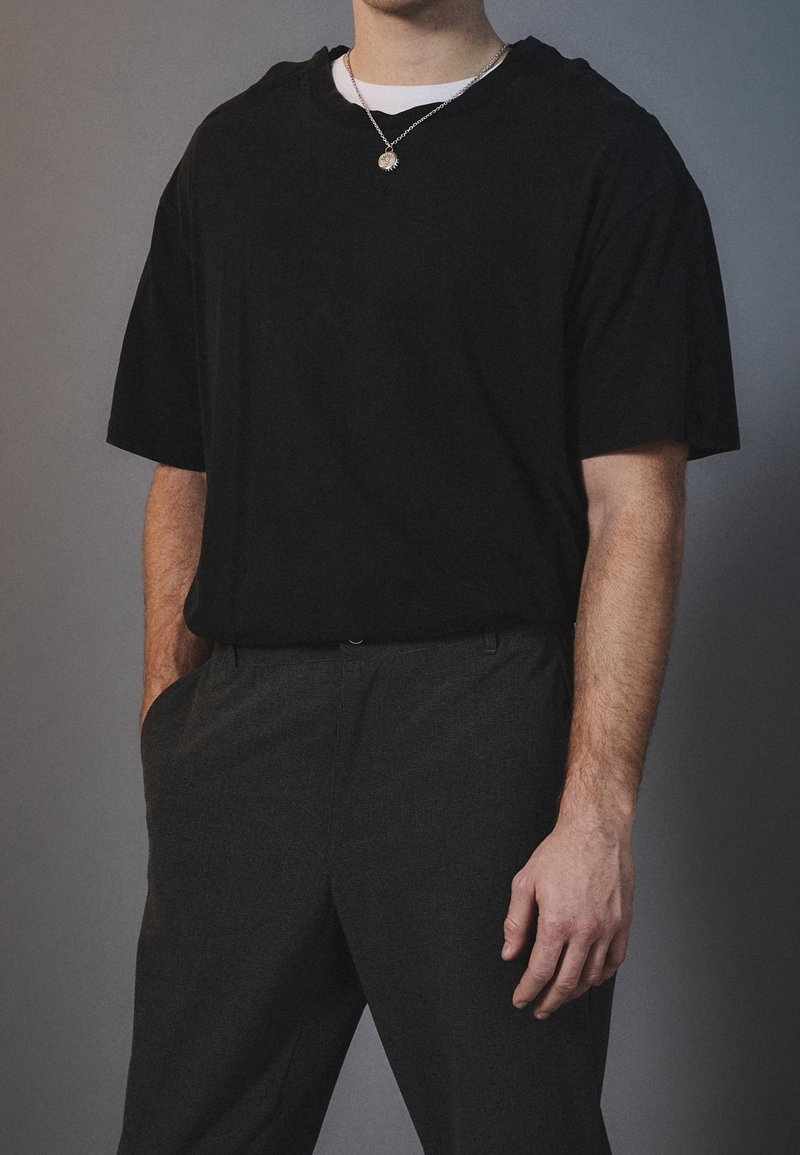 Homme portant un t-shirt noir ample sur un sous-vêtement blanc, un pantalon gris texturé et un collier pendentif argenté sur un fond gris.