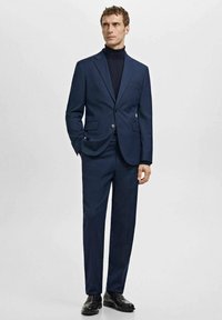 Marineblauer Anzug mit einer eng anliegenden Jacke, klassischem Revers und zwei Fronttaschen, kombiniert mit schwarzen Schuhen und einem dunklen Rollkragenpullover.