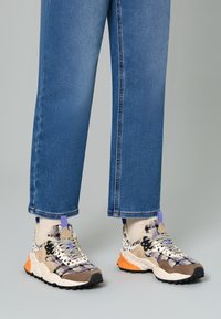 Sneaker multicolori con una tomaia tessurizzata in beige e marrone, accentuate da dettagli viola e arancioni. Abbinati a pantaloni in denim blu.
