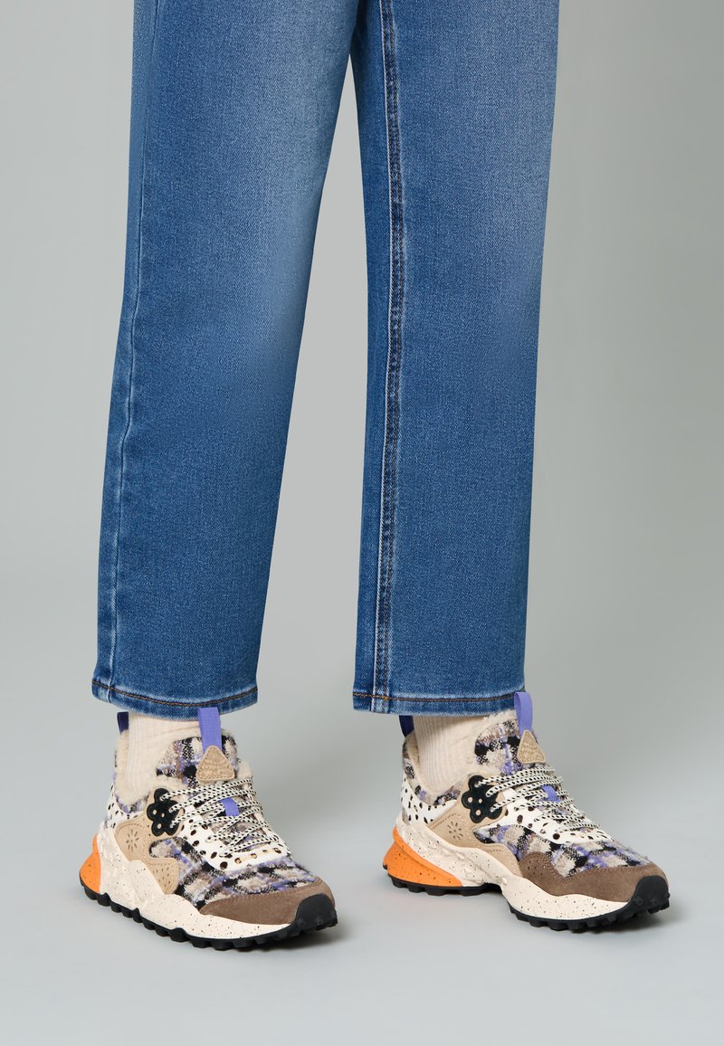 Sneaker multicolori con una tomaia tessurizzata in beige e marrone, accentuate da dettagli viola e arancioni. Abbinati a pantaloni in denim blu.