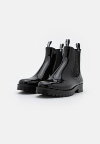 Bottes Chelsea en cuir verni noir avec une texture lisse, semelle en caoutchouc épaisse et languettes de traction. Marquage notable sur les panneaux latéraux élastiques.