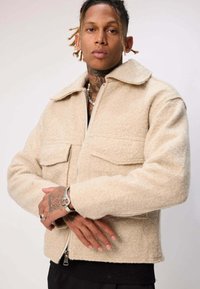 Veste beige en mélange de laine texturée avec une fermeture éclair, un grand col et deux poches poitrine. Présente une coupe courte et des surpiqûres contrastantes.