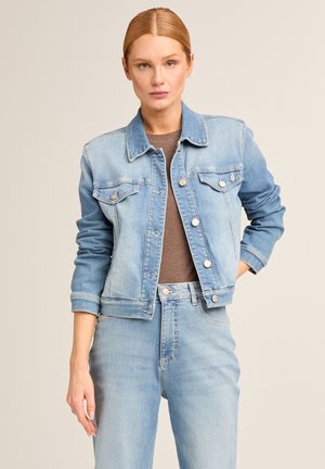 Frau trägt hellblaue Jeansjacke und Jeans mit braunem Hemd, steht mit einer Hand in der Tasche vor neutralem Hintergrund.