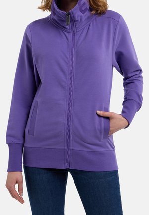 Femme portant une veste violette zippée avec poignets et ourlet côtelés, une main dans la poche, associée à un jean bleu foncé.