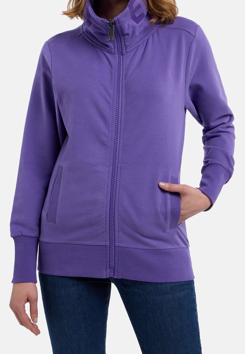 Femme portant une veste violette zippée avec poignets et ourlet côtelés, une main dans la poche, associée à un jean bleu foncé.