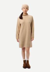 Robe en maille beige à manches longues et coupe décontractée. Associée à un bonnet côtelé assorti. Bottines marron et chaussettes crème visibles.