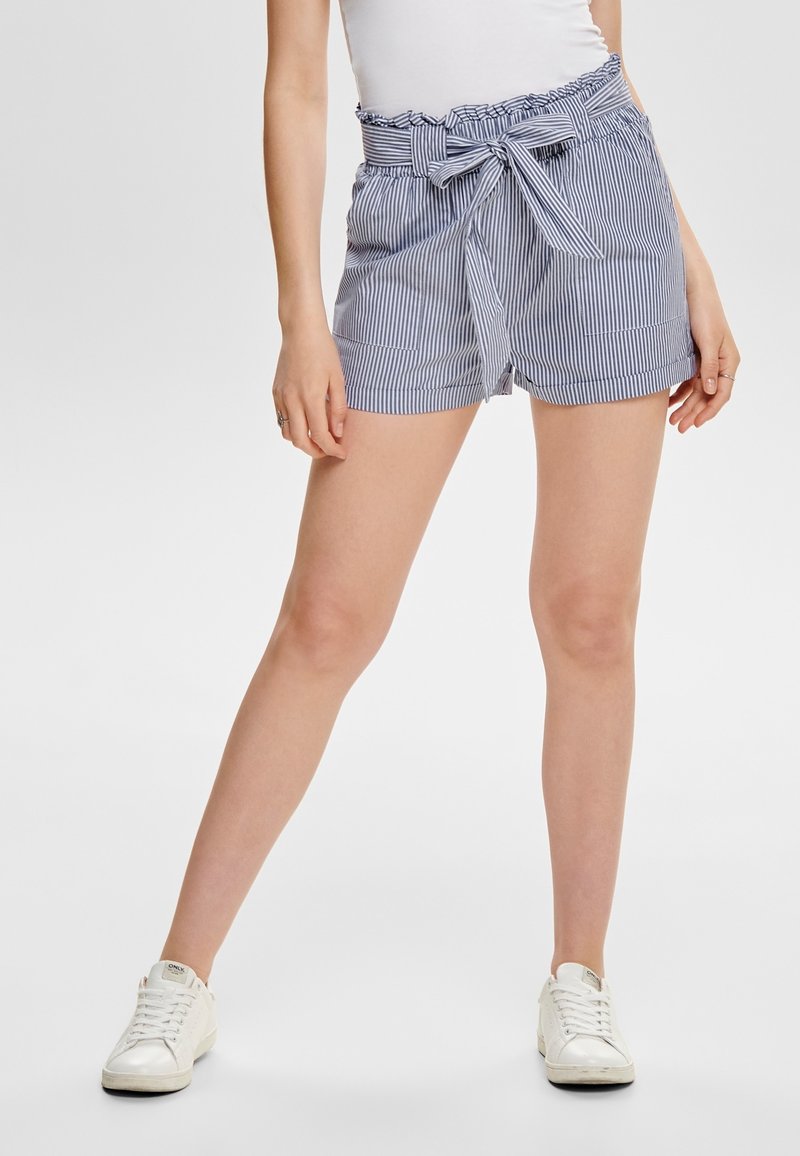 Femme portant des shorts taille haute en papier rayés bleus et blancs avec une ceinture nouée et des baskets blanches, se tenant les jambes écartées.