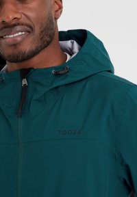 Man wearing teal hooded jacket with zipper and black "TOG24" logo on chest, smiling slightly, against a plain light background.