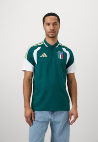 Mann iført grønn og hvit Italia fotball-polo med broderte logoer og lyseblå jeans, står mot en ensfarget hvit bakgrunn.