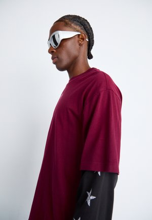 Burgundy oversized t-shirt with short sleeves, paired with a black long-sleeve shirt featuring silver star patterns, and white angular sunglasses.