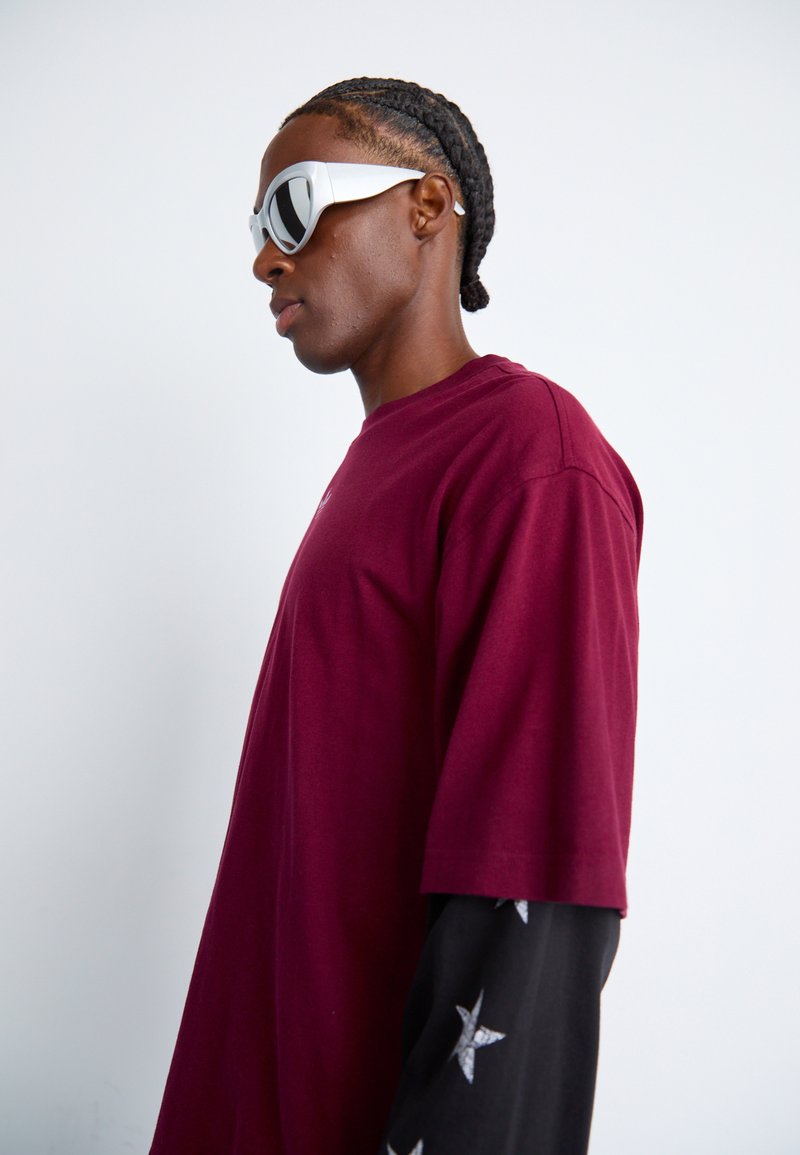 Bordeaux oversized t-shirt met korte mouwen, gecombineerd met een zwarte long-sleeve shirt met zilveren sterpatronen, en witte hoekige zonnebril.