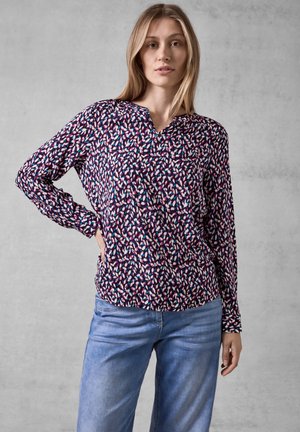 Blusa a maniche lunghe di colore viola con un motivo astratto multicolore. Presenta un collo a V e una vestibilità rilassata. Abbinata a jeans blu.
