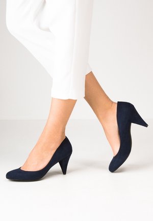 Benen in witte broek, met marineblauwe suède pumps met halvemiddelhoge hak, één voet licht opgetild alsof er gelopen wordt, op een effen lichte achtergrond.