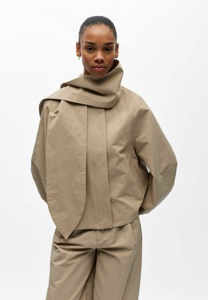 Femme portant une veste beige surdimensionnée à col asymétrique et un pantalon beige assorti, debout les mains dans les poches sur fond blanc.