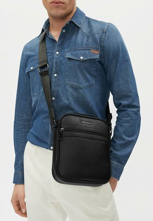 Uomo che indossa una camicia di denim e pantaloni bianchi con una borsa a tracolla nera Gino Rossi con tasca anteriore con cerniera.