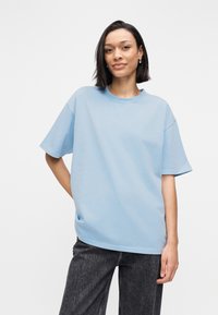 Φαρδιά t-shirt σε ανοιχτό γαλάζιο από μαλακό βαμβάκι, με κοντά μανίκια, στρογγυλή λαιμόκοψη και άνετη εφαρμογή, φορεμένο με σκούρο γκρι τζιν.