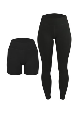 Frau steht barfuß, trägt ein schwarzes Sport-Crop-Top mit weißen Trägern und passende schwarze High-Waist-Bike-Shorts vor einem einfarbigen Hintergrund.