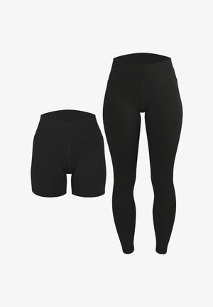Frau steht barfuß, trägt ein schwarzes Sport-Crop-Top mit weißen Trägern und passende schwarze High-Waist-Bike-Shorts vor einem einfarbigen Hintergrund.