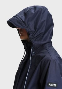 Personne portant une veste de pluie à capuche bleu marine avec une visière et des cordons de serrage ajustables, vue de profil latéral, avec le logo AIGLE sur la manche.