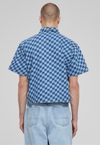 Camicia di jeans a maniche corte a quadri blu con colletto piatto, caratterizzata da un motivo di quadrati blu scuri e chiari e una vestibilità cropped.