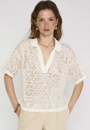 Donna con capelli castano chiaro indossa una polo bianca in pizzo a maniche corte con scollo a V profondo e pantaloni beige, in piedi con le mani in tasca.