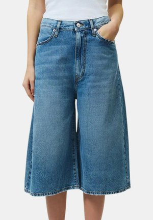 Donna che indossa culotte di denim blu medio a gamba larga con tasche frontali, passanti per cintura e una maglietta bianca infilata dentro.