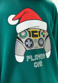 Zeleni pulover s grafikom kontrolera igre koji nosi crvenu božićnu kapu, s natpisom "PLAYER ONE" ispod u pikseliziranom tekstu.