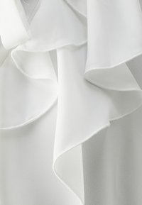 Blusa blanca con volantes en capas. Tejido suave y ligero. Textura lisa con bordes delicados y un sutil brillo. Sin hardware visible.