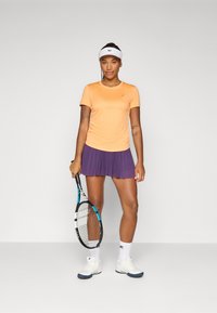 En ung kvinne står selvsikkert og holder et tennisracket. Hun har på seg en oransje skjorte, et lilla plisserte skjørt, og hvite tennissko.