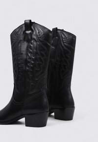 Par de botas de cowboy de cuero negro con costuras decorativas y tacones cuadrados, presentadas sobre un fondo claro y liso.
