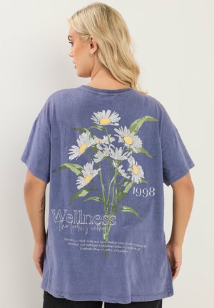 Lyshåret kvinde iført en falmet blå oversized skjorte med hvide margueritter og teksten "Wellness 1998", stående mod en ensfarvet baggrund.