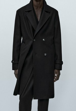 Homme portant un long manteau noir à double boutonnage avec des poignets boutonnés et un pantalon noir assorti sur un fond uni.