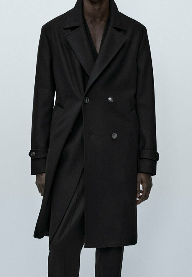 Homme portant un long manteau noir à double boutonnage avec des poignets boutonnés et un pantalon noir assorti sur un fond uni.