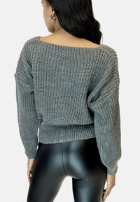 Grauer Strickpullover mit schulterfreiem Design, langen Ärmeln und breitem Bündchen. Kombiniert mit schwarzen, enganliegenden Leggings.