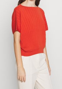Mujer vistiendo un suéter corto de manga, acanalado, de color naranja brillante, combinado con pantalones de alta cintura en un color beige claro, frente a un fondo plano.