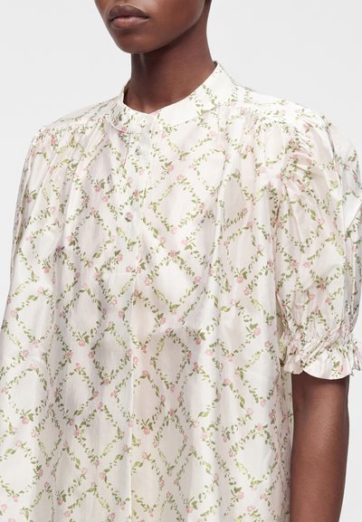 Femme portant un chemisier blanc avec un motif floral en losanges verts et roses et des manches courtes bouffantes avec des poignets froncés.