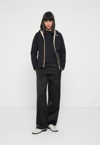 Giubbotto imbottito nero con zip grigia, indossato sopra un top nero; abbinato a pantaloni cargo neri larghi e scarpe bianche.