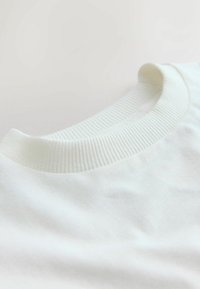 Bela bombažna majica s ribanim okroglim izrezom. Tkanina je gladka z rahlim sijajem, kar poudarja njeno lahko in mehko teksturo.