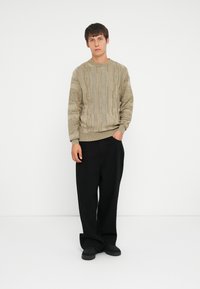 Beige kabelstrik sweater med et tekstureret mønster, parret med sorte bredbenede bukser og sorte slip-on sko. Modellen står foran en hvid baggrund.