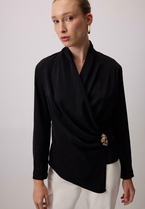 Sort langærmet top med draperet design, asymmetrisk kant og en gylden tekstureret detalje i taljen. Glat stof med en elegant finish.