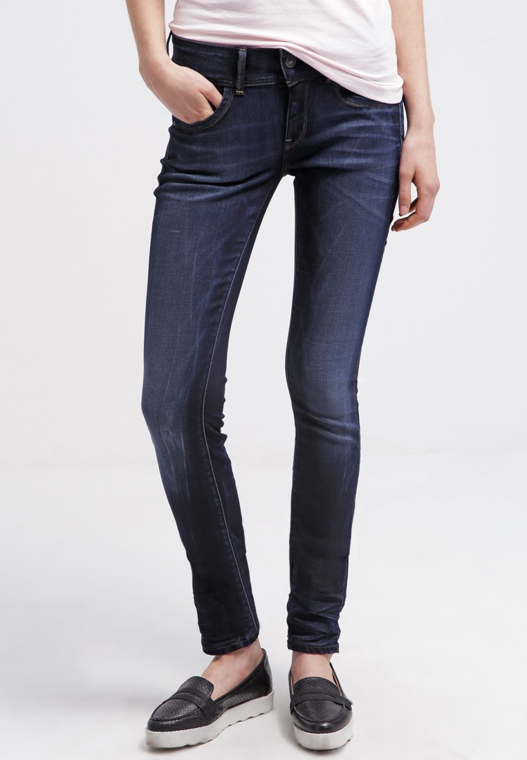 Jeans skinny azul-escuro com ligeiro desbotamento, design de cinco bolsos, fecho frontal com botão e zíper, combinados com sapatos pretos de slip-on.